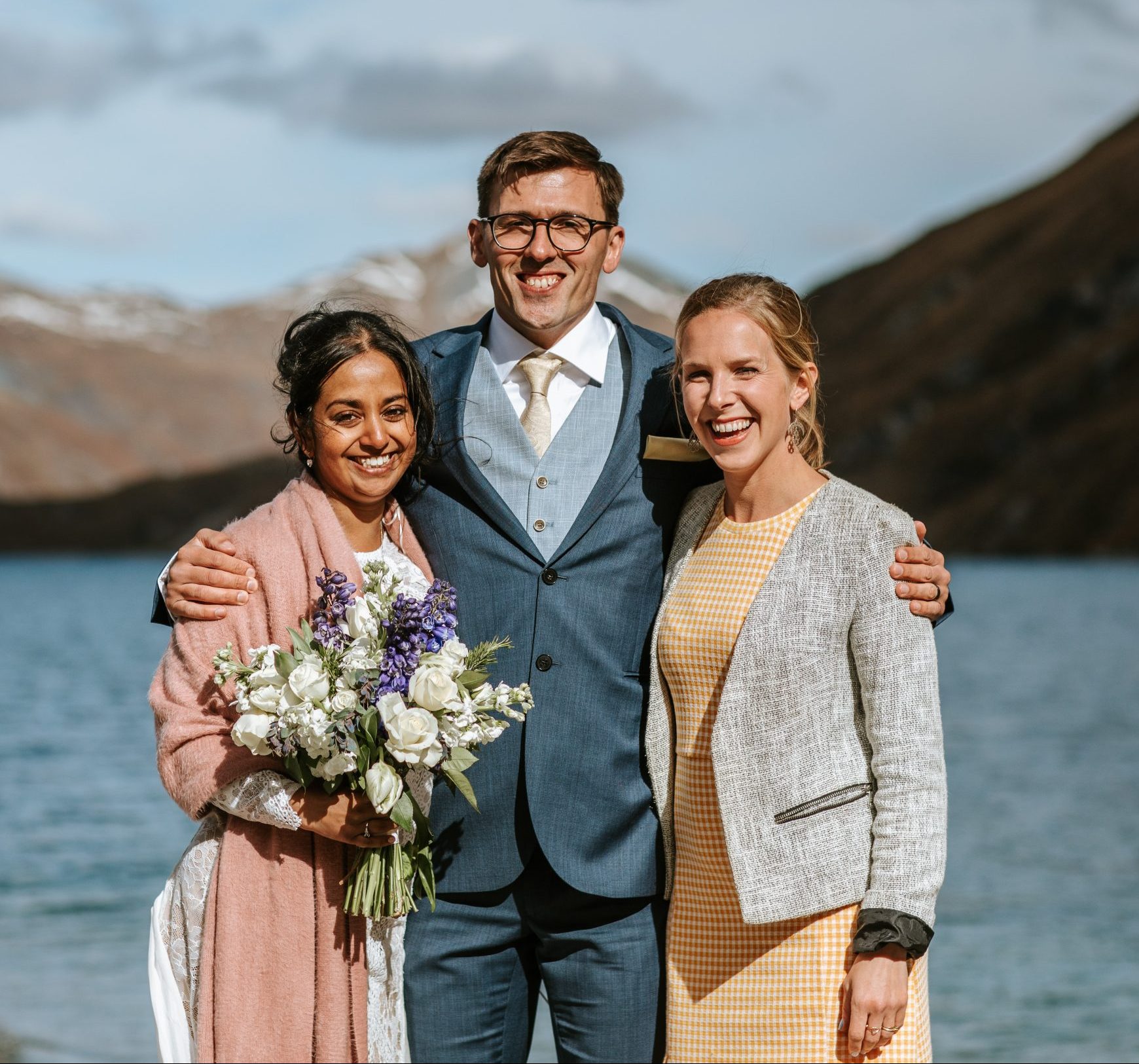 Wanaka helicopter elopment to Lochnagar, post ceremony wedding photo with marriage celebrant Siobhain the Celebrant laughing and smiling with couple with Lochnagar in the background. Bride is holding flowers and draped in a shawl. Part of wedding package with Boutique Weddings NZ