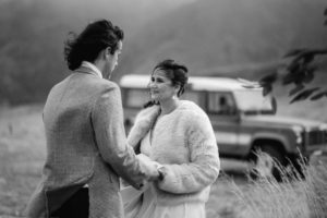 Groom and bride holding hands during ceremony in mountains with retro landrover in the background