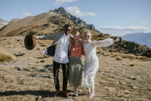 Groom and Bride celebrating with arms out and their celebrant in the middle. Mountain tops in the background