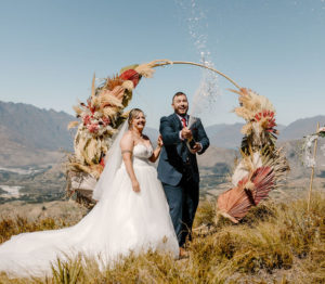 Bride and Groom laugh and spray champagne with mountains in background and flower arch